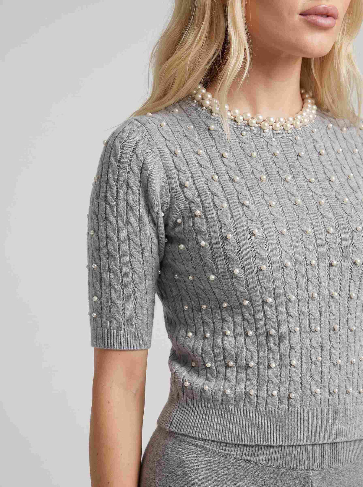 Pulover tricotat gri pentru damă cu perle decorative aplicate pe partea frontală și în jurul gulerului, mâneci scurte și croială confortabilă. Pulover elegant pentru femei, ideal pentru ținute office, casual chic, business și outfituri feminine, ușor de asortat cu jeanși, pantaloni eleganți sau fuste.